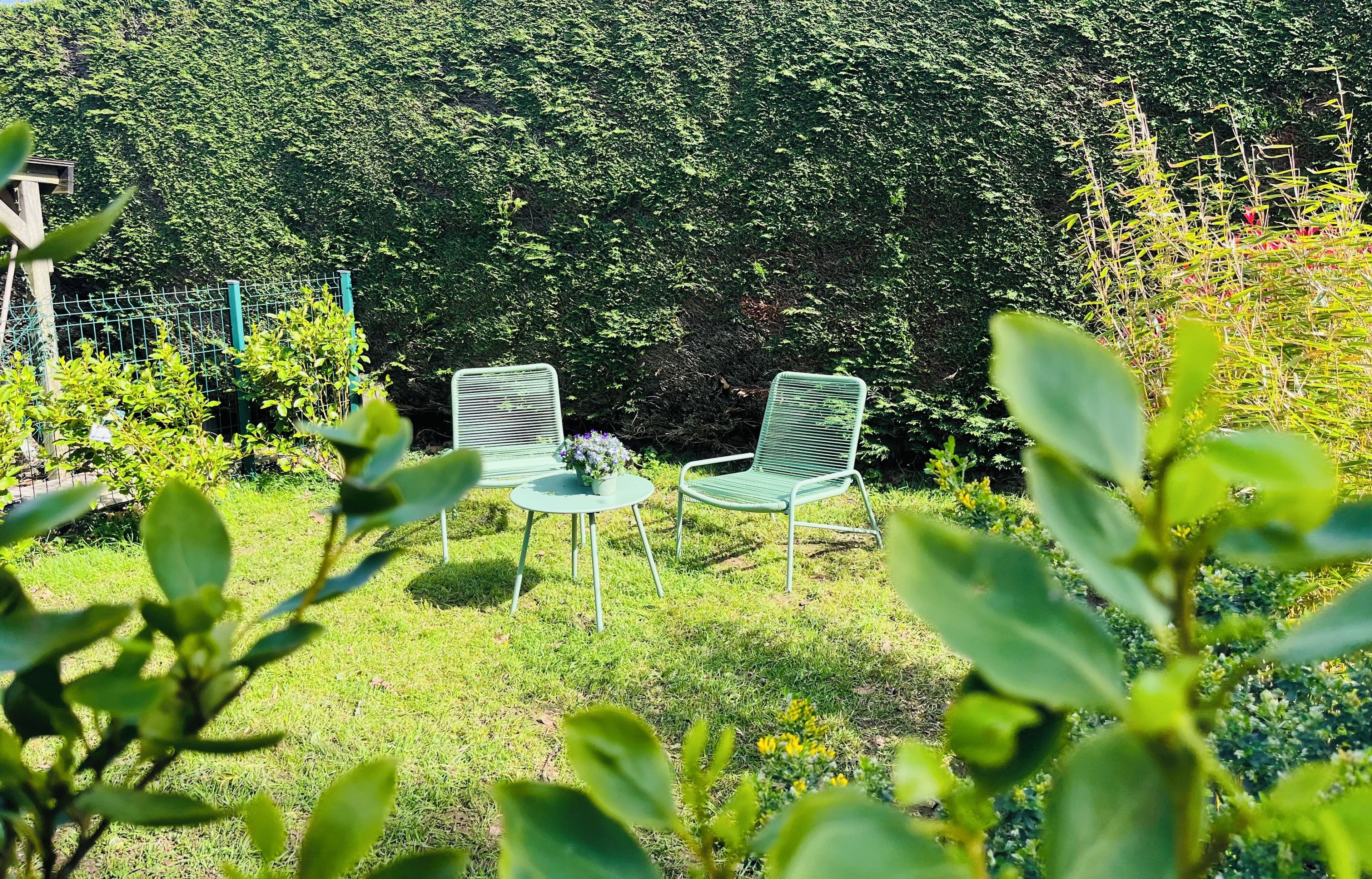Due sedie verdi in metallo con un tavolino e un vaso di fiori in un giardino con un siepe sullo sfondo.