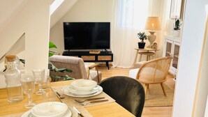 A dining area with a wooden table set for four, a television in the background, and a cozy seating arrangement. - Le Petit Cosy - King Size Bed, Quiet & Parking (Vaucelles)