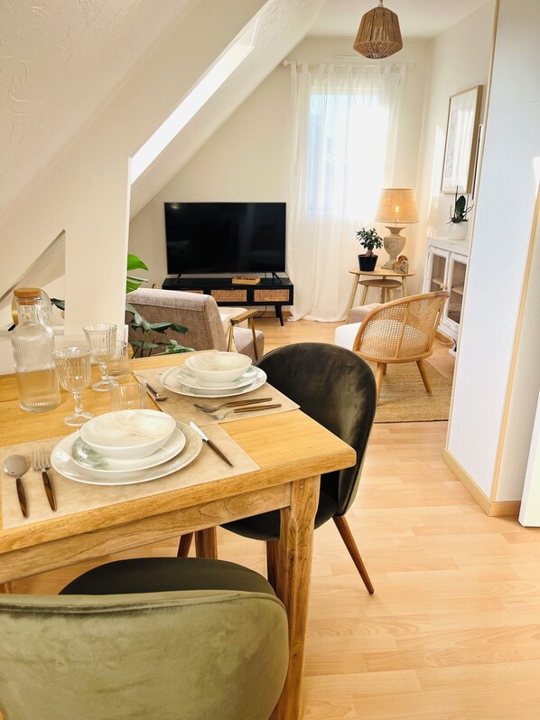 Un coin repas avec une table en bois dressée pour quatre personnes, un téléviseur en arrière-plan et un espace salon confortable.