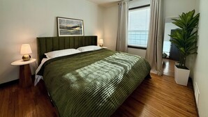 Un dormitorio ordenado con una colcha verde, una lámpara de noche, un cuadro enmarcado en la pared y una planta en maceta.