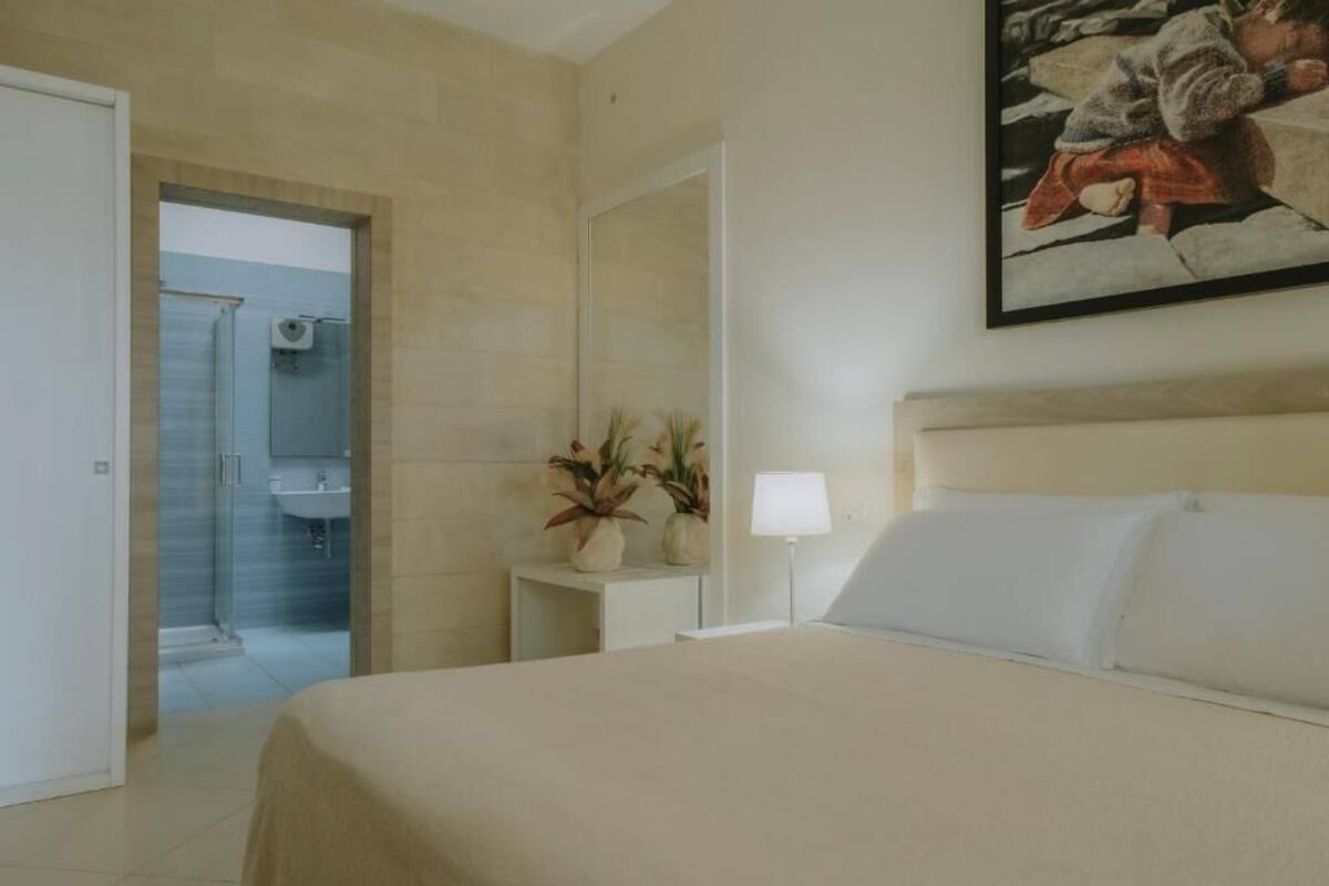 A hotel room with a bed, a bedside table with a lamp, a potted plant, and a painting on the wall.