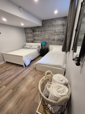 A bedroom with two beds, a nightstand with a lamp, a concrete accent wall, wooden flooring, and a woven basket with towels. - Private 2-brdm basement unit (full bathroomlaundry) (Gatineau)