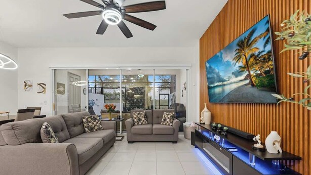 A modern living room with a ceiling fan, a large TV on a wooden wall, a sofa, and a coffee table. - Tranquil Times Townhouse 4 bed Private Pool (Davenport)