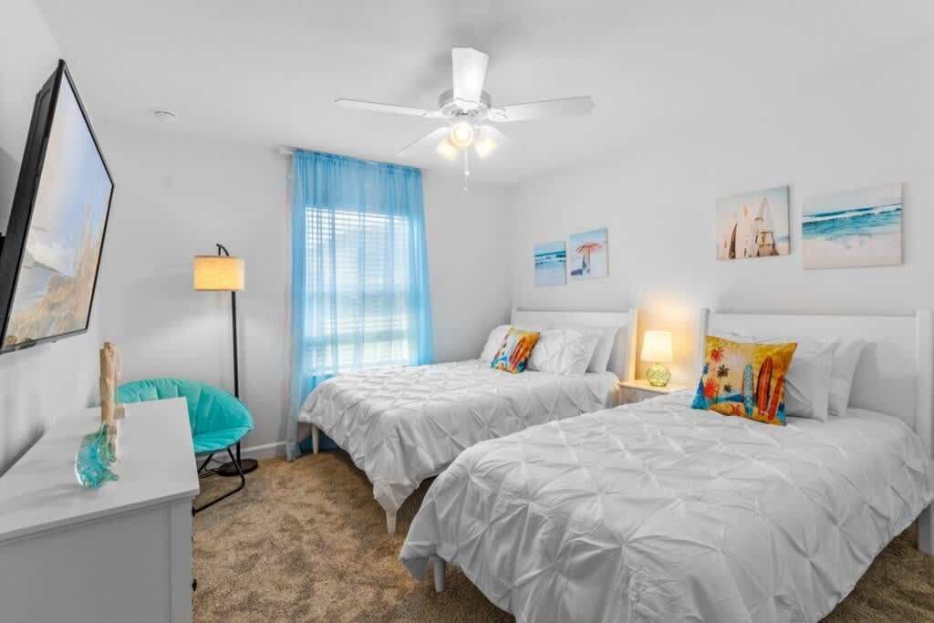 Habitación de hotel con dos camas, un escritorio, una silla, ventilador de techo y obras de arte en las paredes.