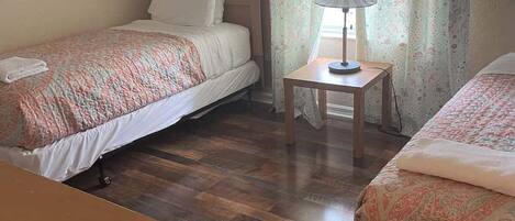 A room with two beds, a wooden floor, a nightstand with a lamp, and a wall sign.