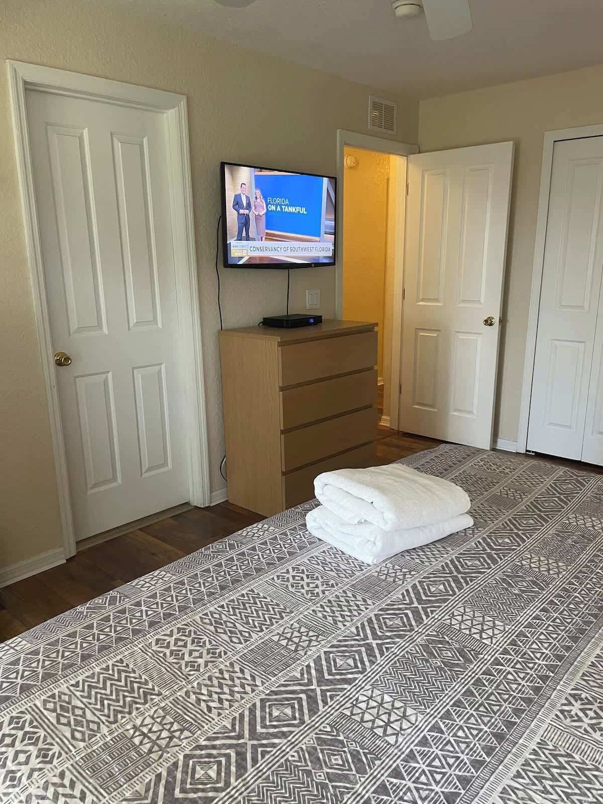 Une chambre d’hôtel avec un lit, un téléviseur fixé au mur, une commode et un tapis à motifs.