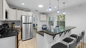 Cozinha moderna com armários brancos, geladeira de aço inoxidável, ilha de vidro com banquetas, iluminação pendente e janela com persianas.