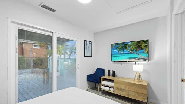 Quarto com cama, televisão fixada na parede, cómoda de madeira, cadeira azul e porta de vidro deslizante para área externa.