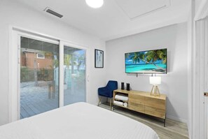 A bedroom with a bed, a TV mounted on the wall, a wooden dresser, a blue chair, and a sliding glass door leading to an outdoor area. - Palm Cove Hideaway 2bd Close to the Beach (Madeira Beach)