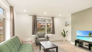 A living room with a green sofa, a grey armchair, a coffee table, a flat-screen TV, and a view of a residential area through large windows. - 3bed House - Close to Worksop Mansfield (Mansfield)