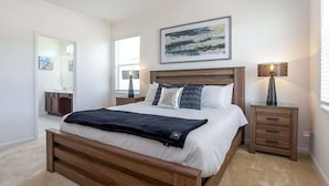 Un dormitorio con una cama de madera, una mesita de noche con lámpara, un cuadro en la pared y un baño visible a través de una puerta abierta.