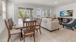 A modern living room with a dining area, a sofa, a TV, and a large window with blinds. - Beautiful 6br5bathpool Clubhouse (Davenport)
