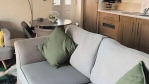 A modern living room with a grey sofa, a yellow pillow, and a green pillow. There is a dining table with a glass bowl and a potted plant. The room has wooden cabinets and a tiled floor. - 1-bed Hideout in Ripon With Free Parking (Ripon)