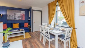 A dining area with a white table set for four, a blue sofa with orange cushions, and a cityscape painting on the wall. - 1 Bedroom Scottish Cottage-on Street Parking (Selkirk)