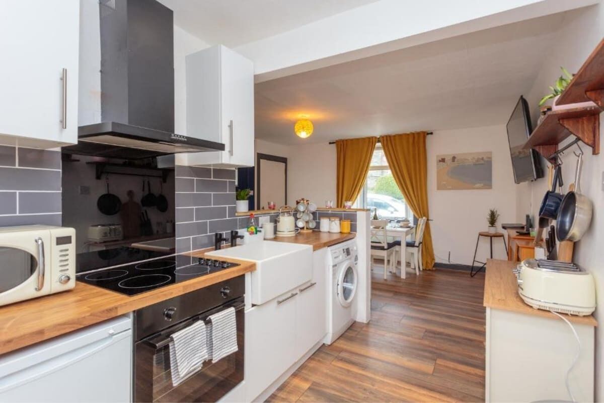 Ett modernt kök med mikrovågsugn, spis, ugn och tvättmaskin. Det finns en matplats med ett bord och stolar, och en tv som är monterad på väggen.