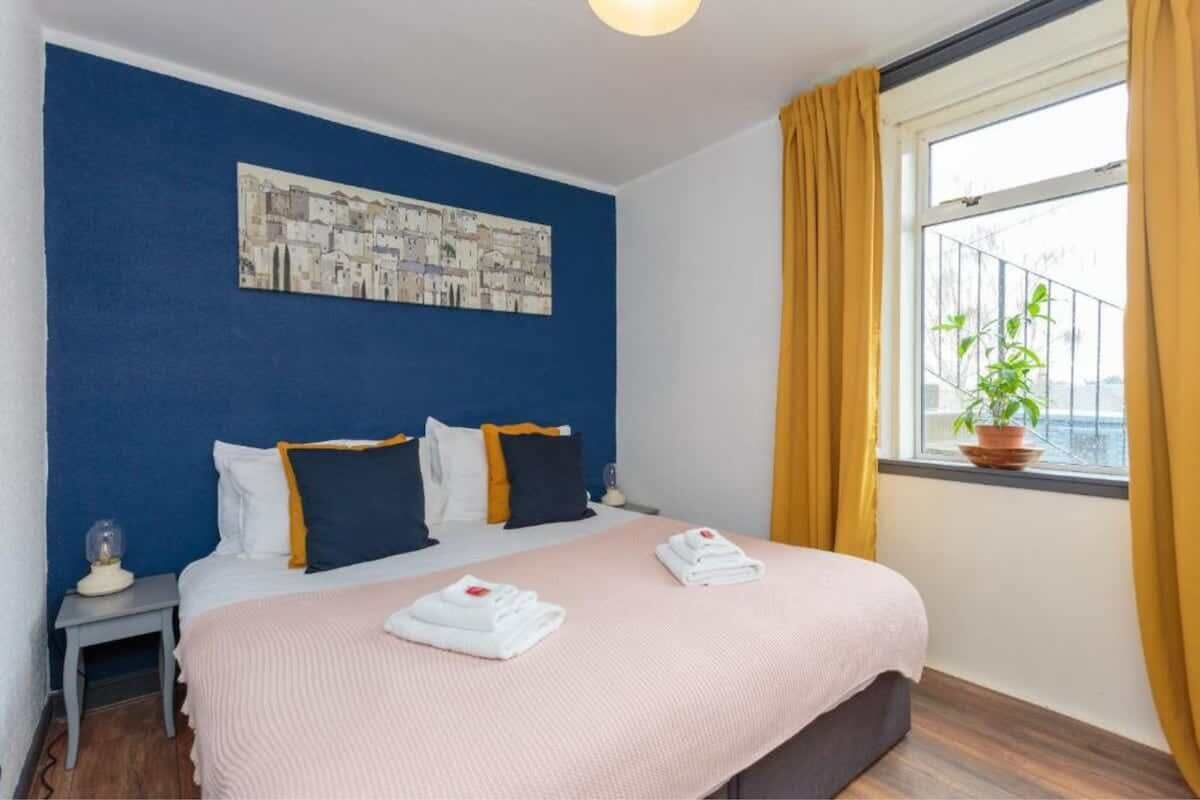 A hotel room with a bed, a nightstand, a window with curtains, a potted plant, and a cityscape painting on the wall.