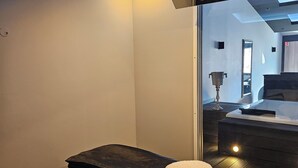 A modern bedroom with a large bed, a massage table, and a mirror reflecting a bathroom with a bathtub and a vanity. - Love Room Le Jardin D'Éden (La Motte-Servolex)
