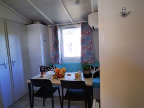 A compact kitchen with a dining table, chairs, a window with curtains, and a built-in bench. - Cozy Mobile-home with Terrace for 5 people (Villecroze)
