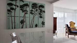 Ein moderner Essbereich mit einem Glastisch und Stühlen, einem großen Baumgemälde an der Wand und Blick ins Freie durch leichte Vorhänge.