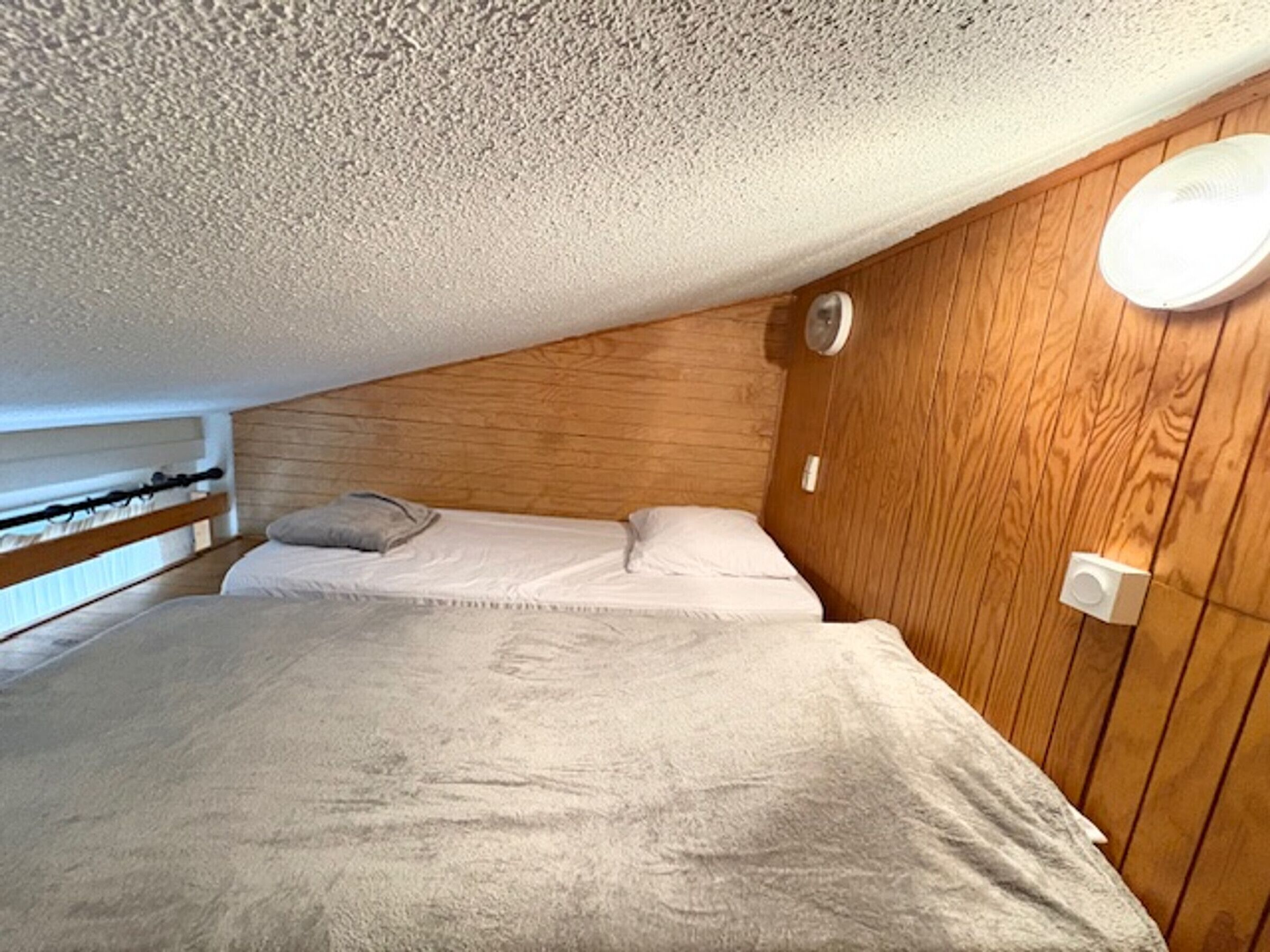 A small, cozy room with a sloped ceiling, wooden panel walls, a bed with white linens, and a wall-mounted light fixture.