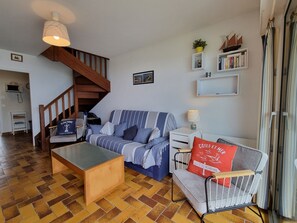 Un salón con una mesa de centro de madera, sofá a rayas y un sillón gris con un cojín rojo. Se observa una escalera con barandillas de madera al fondo.
