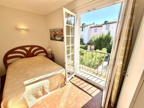 Ein Schlafzimmer mit Bett, Nachttisch, Wandleuchte, einem Gemälde und einem Balkon mit Blick auf Gebäude und Grünflächen.
