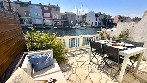 Eine Terrasse mit Steinboden, einem Tisch mit Stühlen, einer Couch mit Kissen und Blick auf einen Hafen mit Gebäuden und Booten.