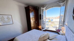 A bedroom with a bed, a window with a view of buildings, a wooden wardrobe, and a framed picture on the wall. - 3-bedroom apartment with sea view, beach access, parking, in Port Grimaud (Port Grimaud)