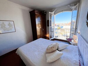 A bedroom with a bed, a window with a view of buildings, a wooden wardrobe, and a framed picture on the wall. - 3-bedroom apartment with sea view, beach access, parking, in Port Grimaud (Port Grimaud)