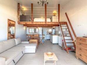 Ein großzügiger Wohnbereich mit hohen Decken, Holzbalken, einem Esstisch, einer Couch, einem kleinen Tisch mit einer Schale und einer Treppe, die zu einer oberen Ebene führt.