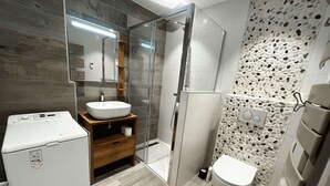 Une salle de bain moderne dotée d’un lave-linge encastré, d’une cabine de douche en verre et de toilettes avec un mur décoré d’un motif de galets.