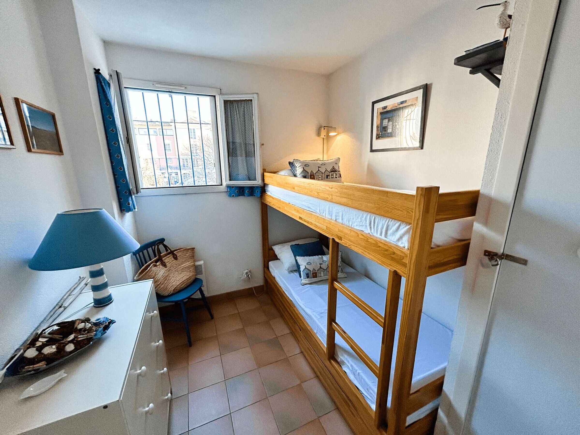 Una habitación pequeña y ordenada con una cama individual superpuesta, un escritorio con lámpara, una silla y una ventana con cortinas.