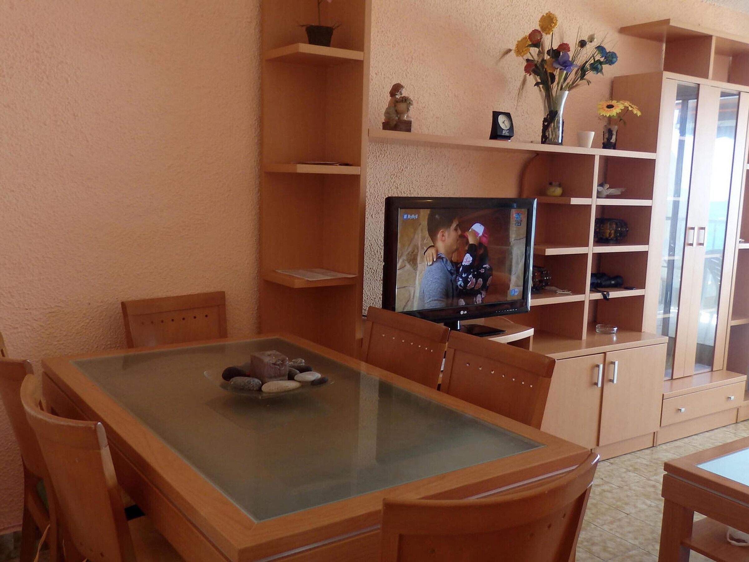 Een woonkamer met een eettafel met glazen blad, houten stoelen, een televisie en een boekenkast met decoratieve objecten.