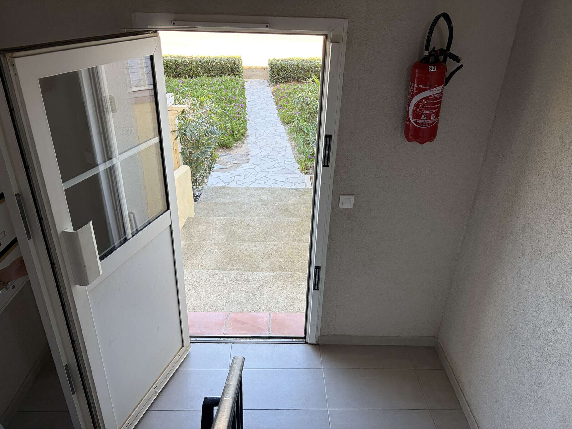 Un couloir avec une porte donnant sur un jardin, un extincteur fixé au mur et une rambarde métallique au premier plan.