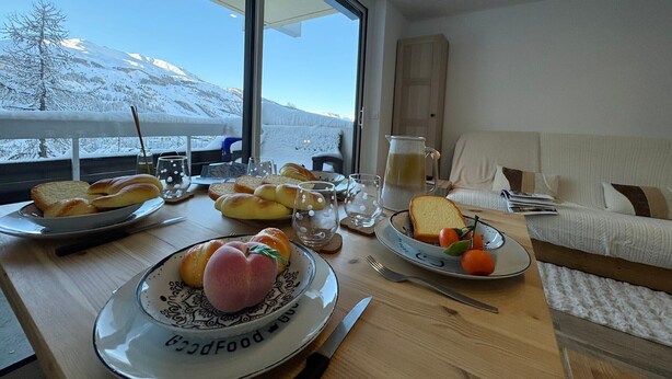 A breakfast table with bread, pastries, and fruit, a pitcher, and a view of snow-covered mountains through large windows. - Vars - Cozy Apartment for 4 People (Vars)