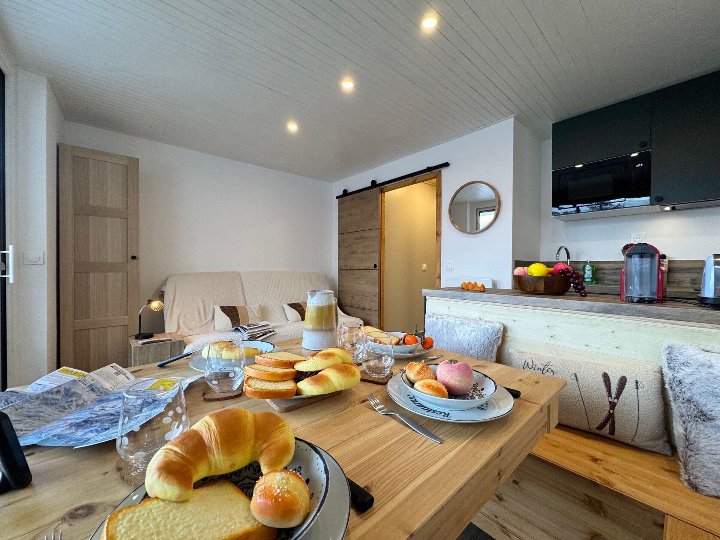A cozy living room with a wooden dining table set for breakfast, a sofa, and a kitchen area with modern appliances.