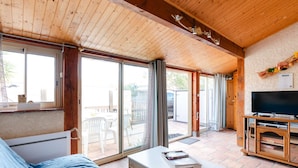 Una habitación acogedora con techo de madera, un sofá, un televisor y una puerta de vidrio corredera que da a un área exterior con un patio y sillas.