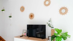 A modern living room with a flat-screen TV on a wooden cabinet, surrounded by white walls and decorative sunburst wall art. - 3-room Pavilion with sea view, pool and terrace, pets welcome (Fleury-Saint-Pierre-la-Mer)