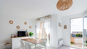 A bright, modern kitchen with a dining area, white cabinetry, a TV, and a large window. - 3-room Pavilion with sea view, pool and terrace, pets welcome (Fleury-Saint-Pierre-la-Mer)
