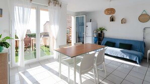 A bright, sunlit room with a sliding glass door leading to a patio, a dining table with chairs, a blue sofa, and a small refrigerator. - 3-room Pavilion with sea view, pool and terrace, pets welcome (Fleury-Saint-Pierre-la-Mer)