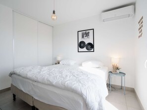 A modern bedroom with a large bed, bedside lamps, a framed picture, and an air conditioning unit. - Bright, air-conditioned apartment with WIFI and parking in Narbonne-Plage (Narbonne-Narbonne Plage-Narbonne Plage)