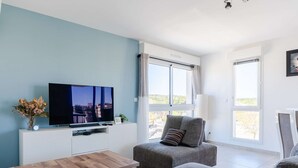 Ein modernes Wohnzimmer mit Flachbildfernseher, grauen Polstermöbeln, einem Couchtisch aus Holz und großen Fenstern.