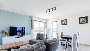 Ein modernes Wohnzimmer mit einem Flachbildfernseher, grauen Sofas, einem Holztisch, einem Esstisch mit Stühlen und großen Fenstern mit Vorhängen.