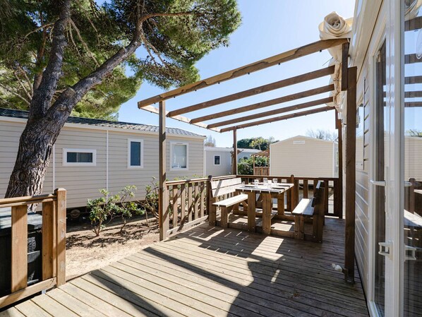 Une terrasse en bois, agrémentée de tables de pique-nique et de bancs, entourée d’une pergola en bois et jouxtant une maison mobile.