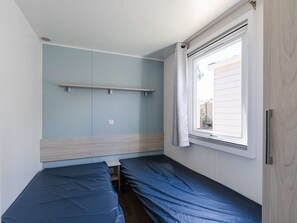 Une petite pièce avec deux lits, une fenêtre avec des rideaux, une étagère et un interrupteur.