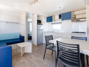 A compact kitchen with a refrigerator, stove, and microwave. There is a dining area with a table and chairs. - Comfort mobile home 450 m from the sea, 6 ppl (Narbonne)