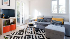 Een moderne woonkamer met een grijze bank, gele kussens, een zwart-wit vloerkleed, een tv op een houten dressoir, en een raam met uitzicht op groen.