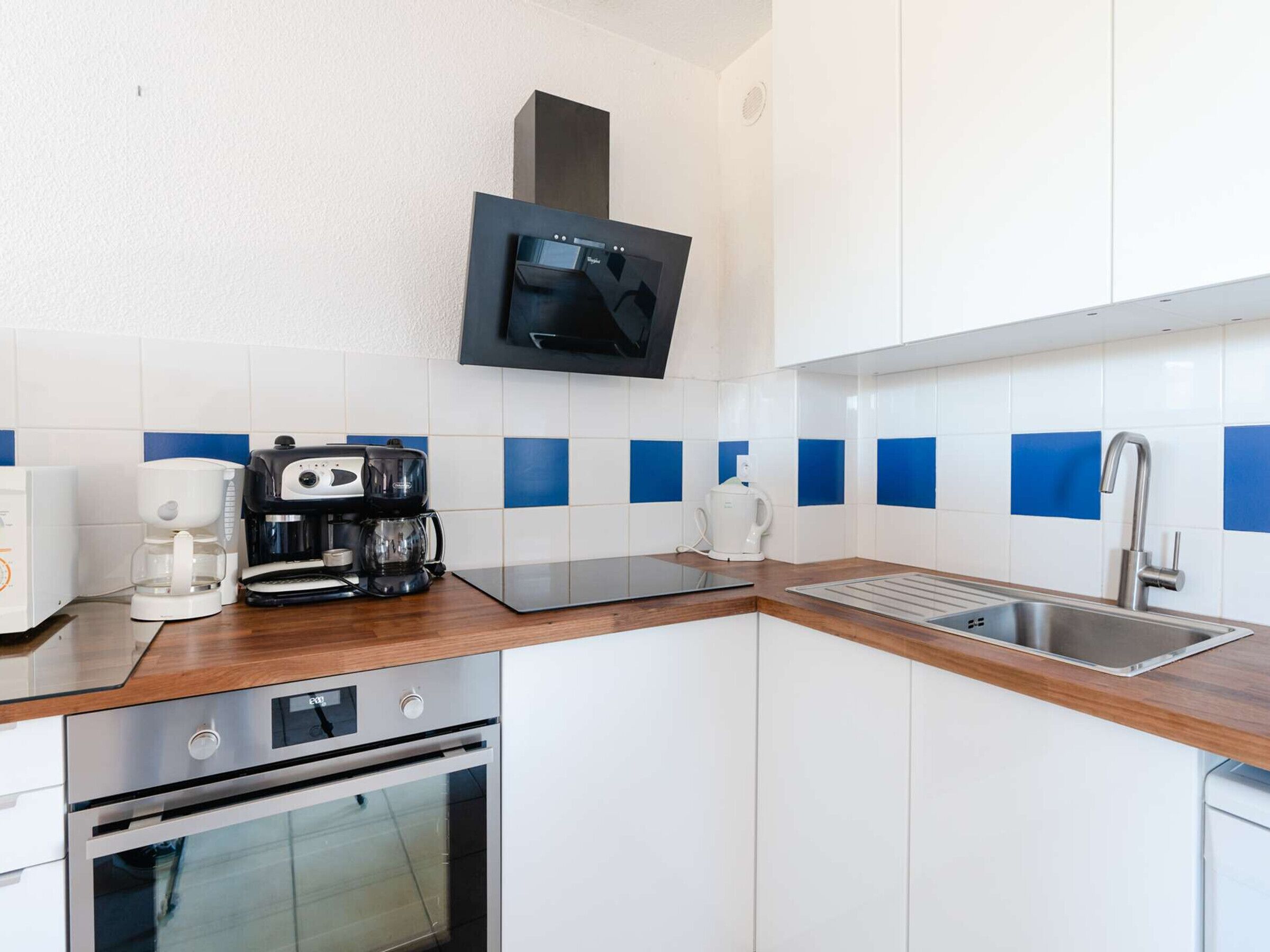 Een moderne keuken met witte kasten, blauwe tegels, een roestvrijstalen spoelbak, een koffiezetapparaat, een magnetron en een inbouwoven.