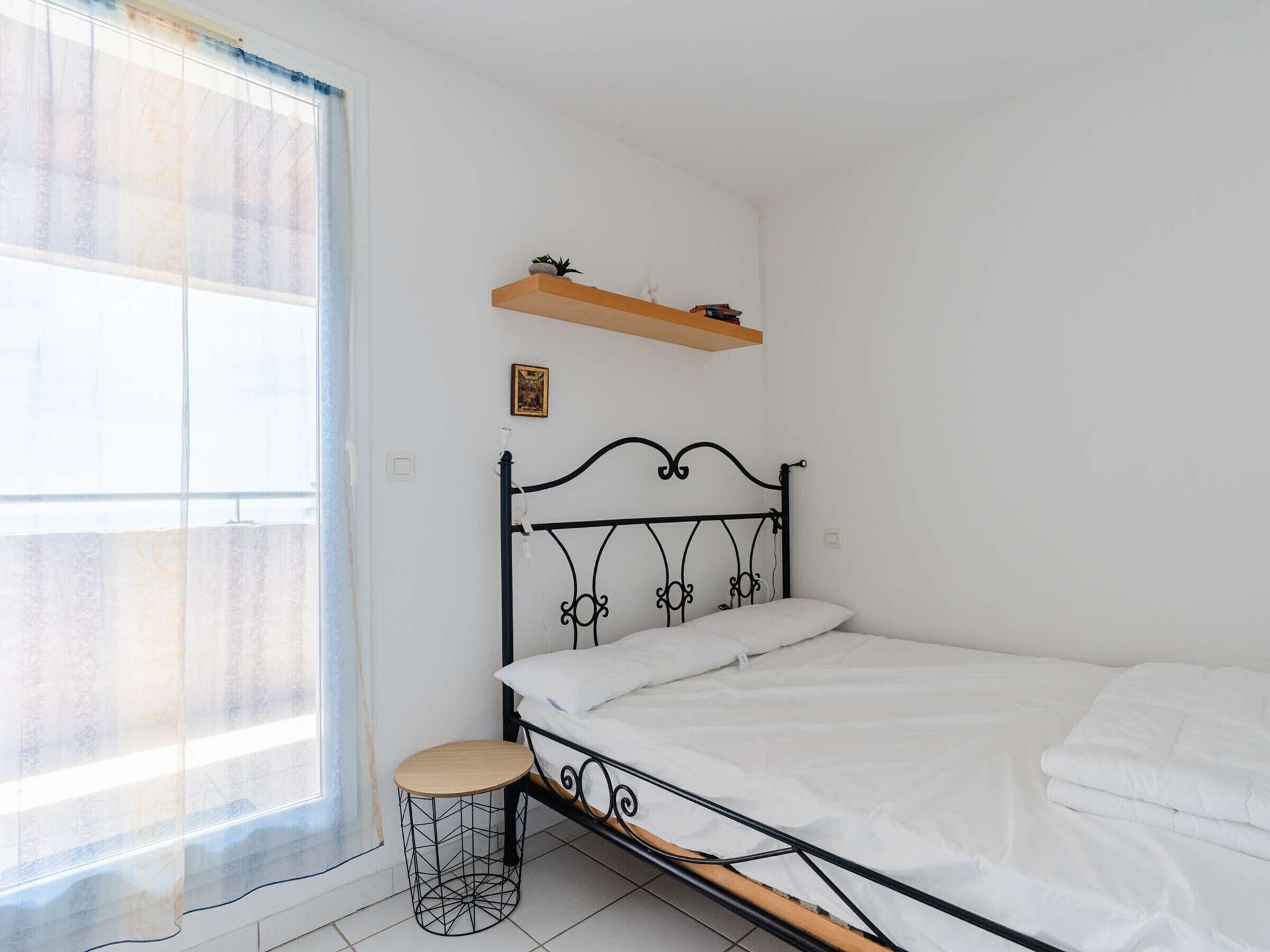 A bedroom with a metal bed frame, a small round stool, a window with sheer curtains, and a wooden shelf above the bed.