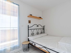 Ein Schlafzimmer mit einem Metallbettgestell, einem kleinen runden Hocker, einem Fenster mit leichtem Vorhang und einem Holzregal über dem Bett.